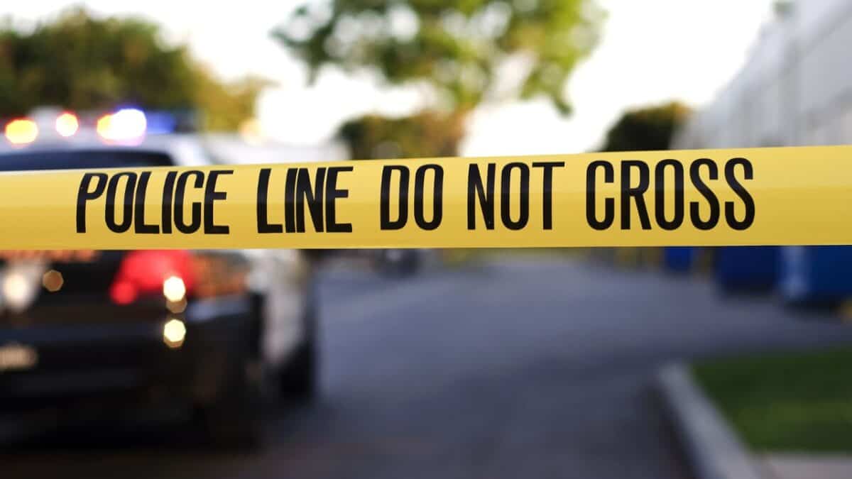 Close-up of a 'POLICE LINE DO NOT CROSS' tape with a blurred patrol car and its flashing lights in the background.