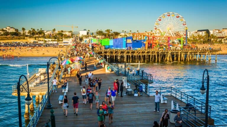 A bustling pier with crowds of people, a Ferris wheel, and amusement rides, overlooking a sunny beachfront.