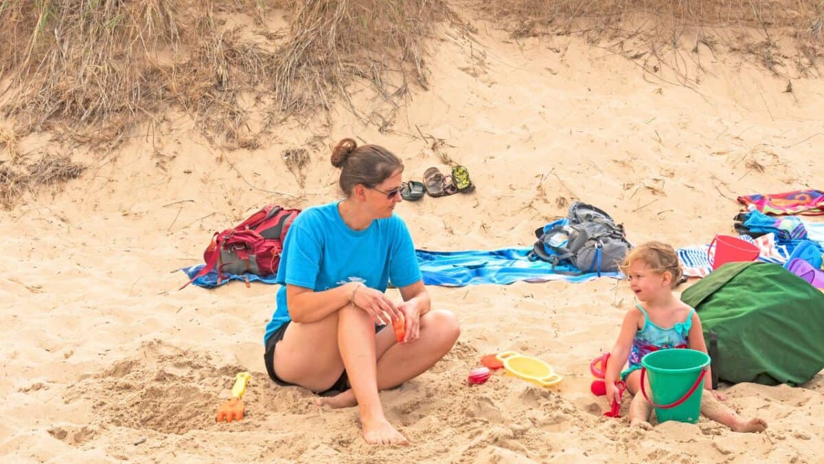 A woman and a young child sitting on a beach with sand toys, surrounded by bags and towels, with dune grass in the background.