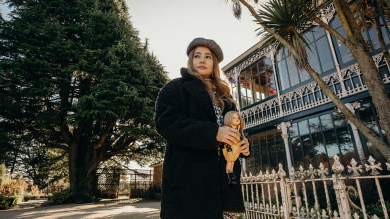 A woman in a black coat and beret holds a doll, standing outside Larnach Castle, New Zealand on a sunny day, with tall trees in the background.