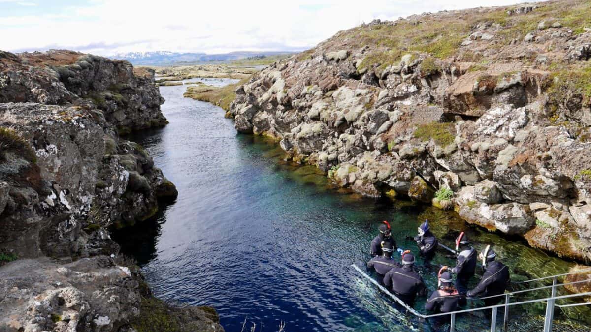 Divers in wetsuits preparing to enter a clear blue fissure between rocky cliffs in a rugged landscape.