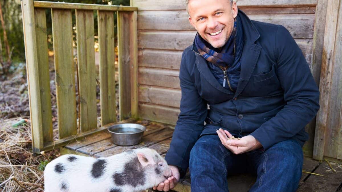 A smiling man crouched down next to a small piglet inside a wooden pen.