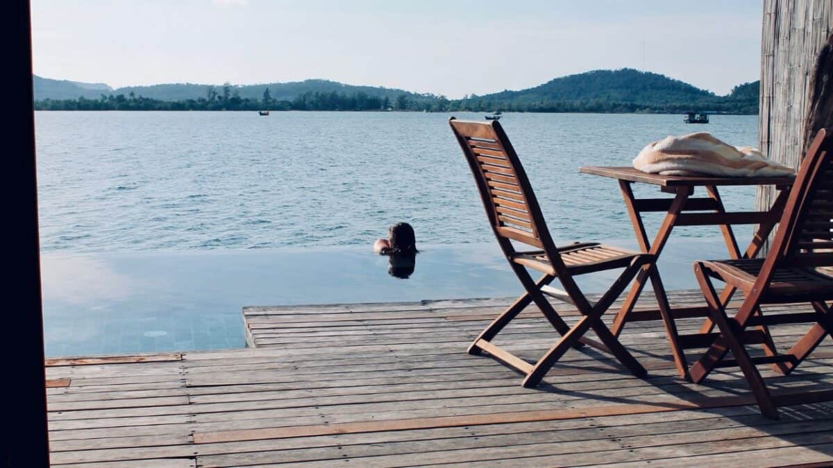 A person enjoying a swim in a serene, infinity-edge pool overlooking a lake, with wooden chairs and a table on the deck in the foreground.