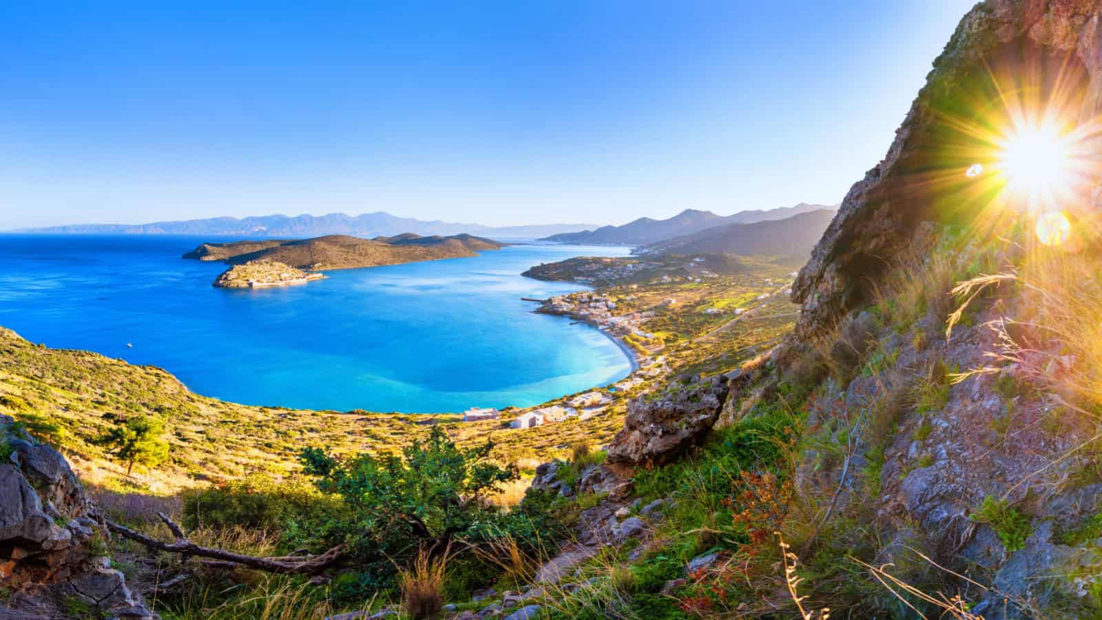 Best Greek Island for Romantic Couples 13 A panoramic view of a coastal landscape at sunrise, featuring a sunburst effect beside rugged mountains, overlooking a serene bay with clear blue water and distant islands.