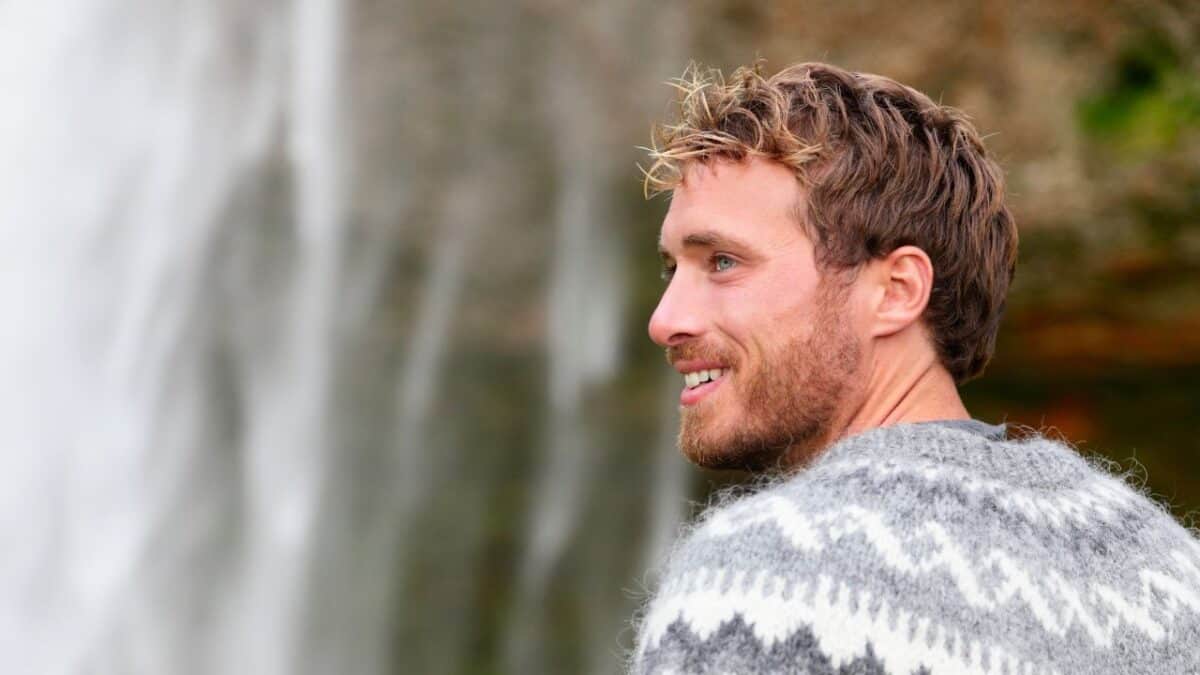 A man in a traditional Icelandic sweater smiling and looking away with a waterfall in the soft-focused background.