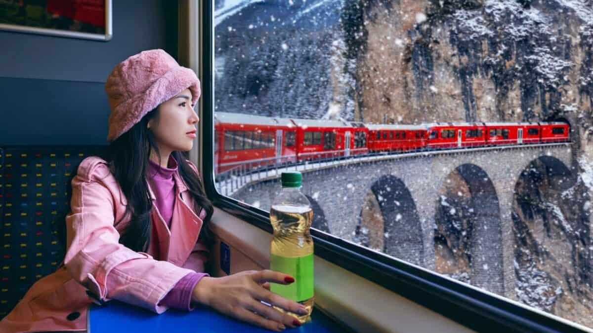 A woman in a pink coat and beret looking out the window at a red train traveling through a snowy landscape.