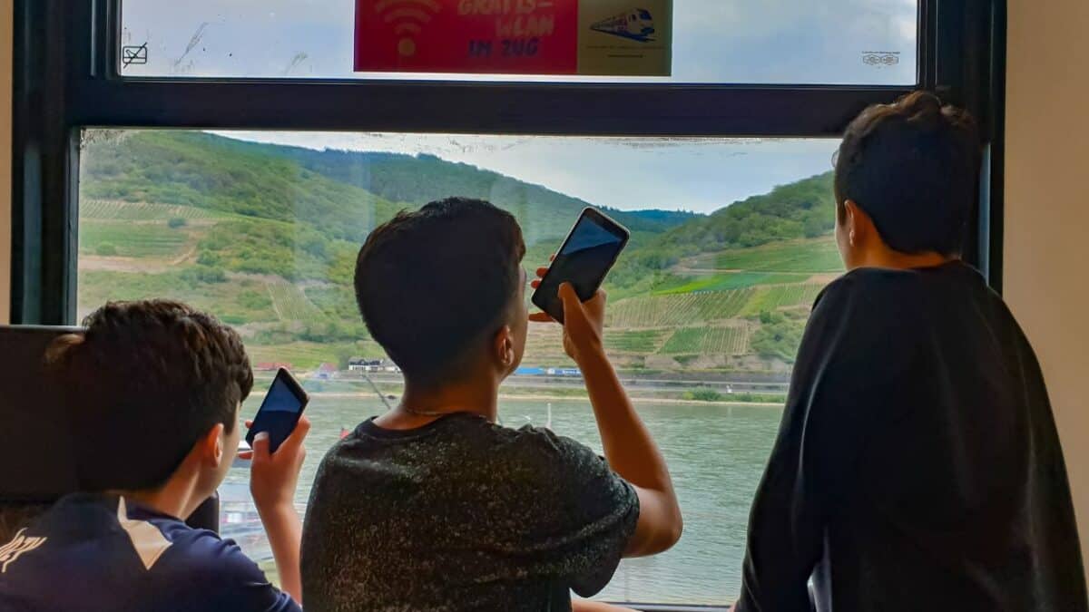 Three children inside a train using their mobile devices by the window with a view of hills and a river outside.