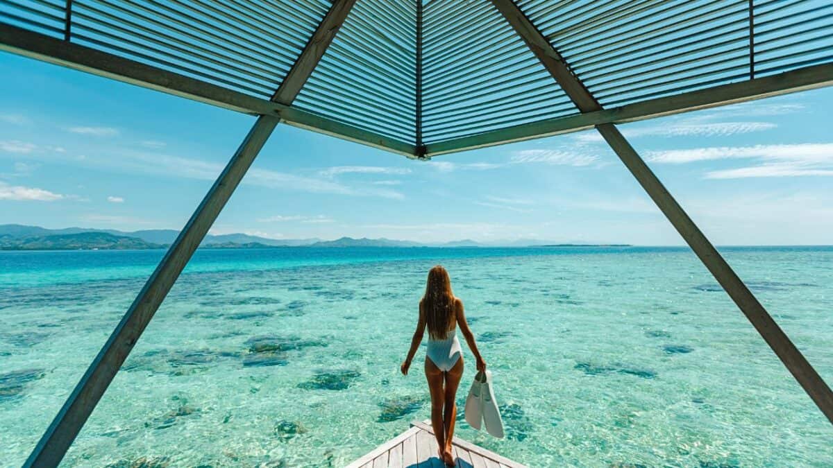 A person with long hair in a white swimsuit holding a pair of flippers, standing at the edge of a wooden pier with a louvered roof, looking out over a clear turquoise sea.