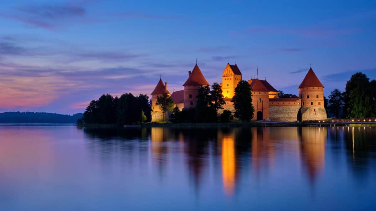 A serene evening view of a medieval castle with illuminated walls and turrets reflected in the calm lake waters during dusk.