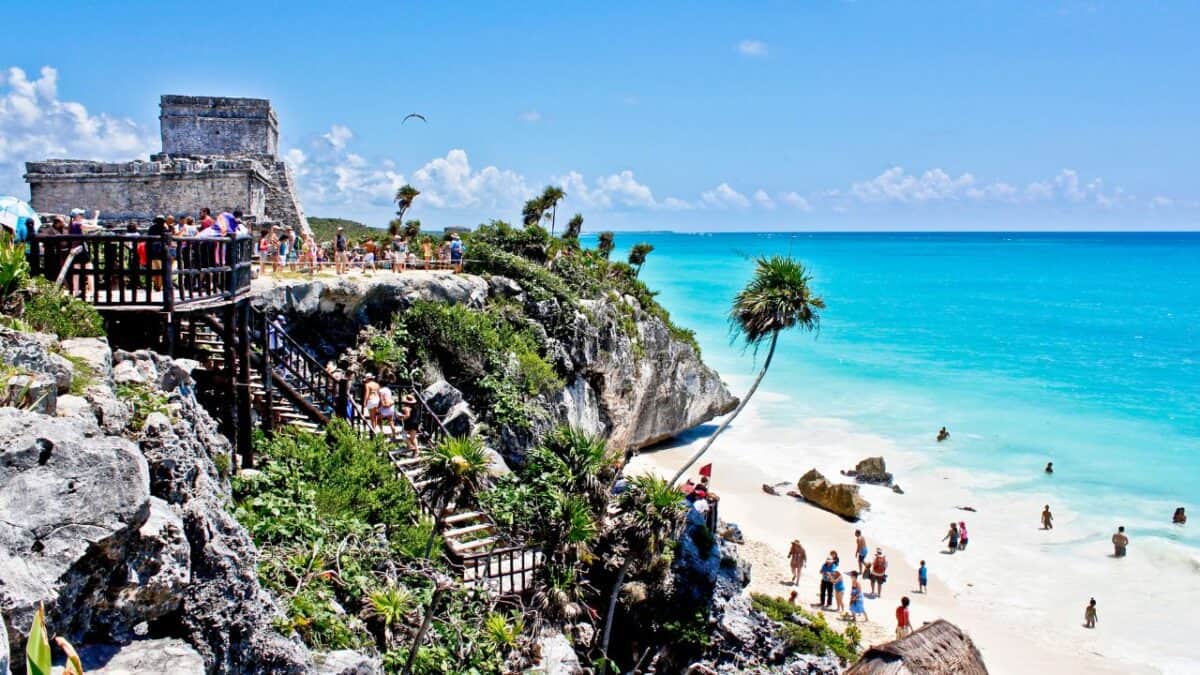 A vibrant beach scene with visitors exploring ancient ruins atop a cliff overlooking a turquoise sea, palm trees, and sandy shores below.