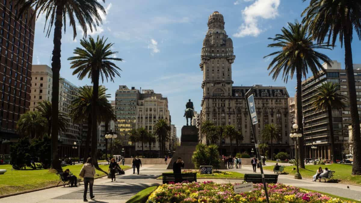 A vibrant city park scene with people walking and sitting, flanked by tall palm trees, colorful flower beds, and a grand historic tower under a clear blue sky. a statue on a pedestal centers the image.