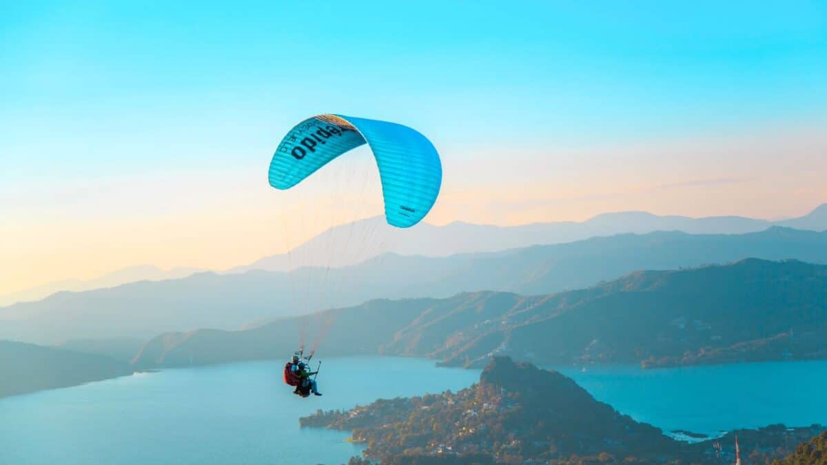 A paraglider floats in the sky above a scenic landscape of mountains and a lake during sunset, with warm golden light bathing the scene.