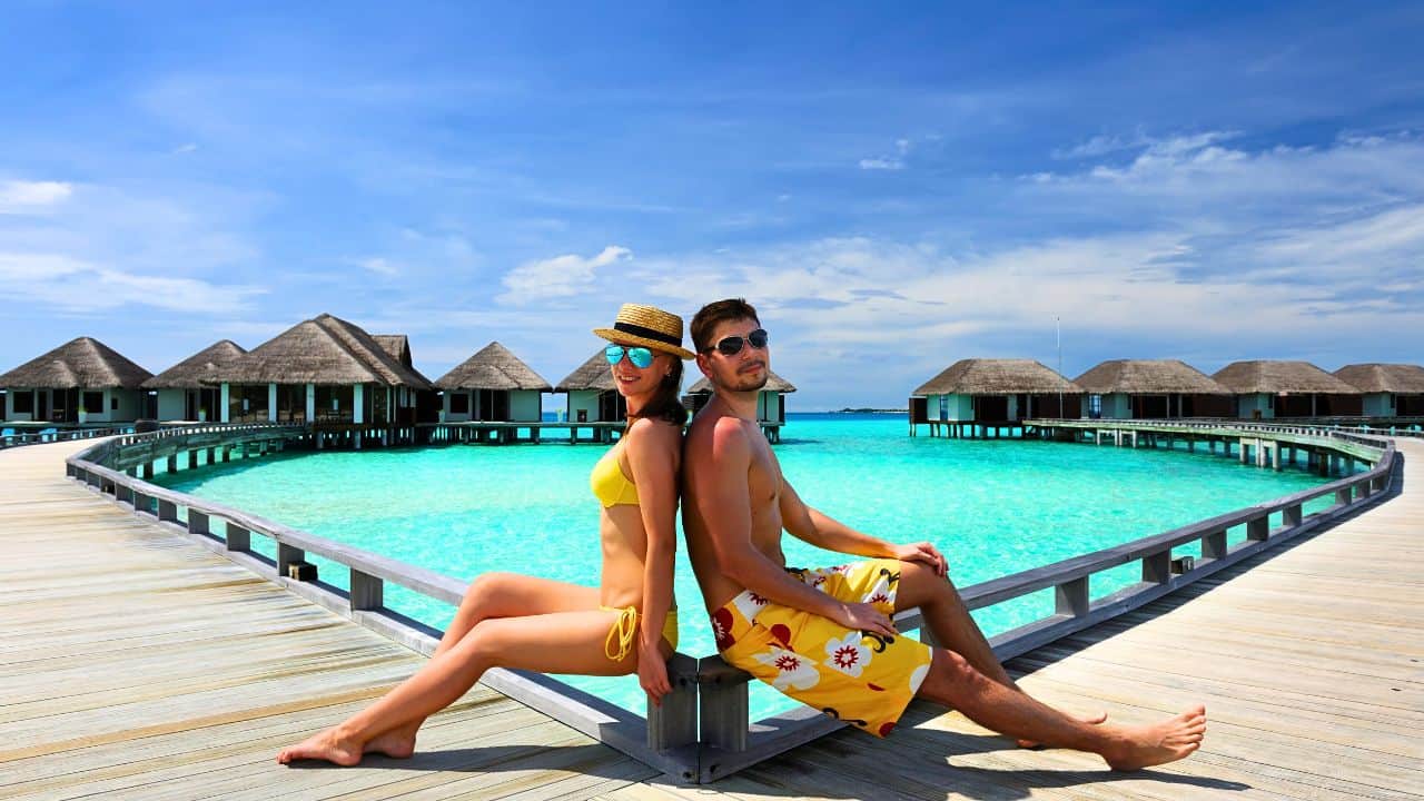 A couple sitting back-to-back on a wooden pier over clear turquoise waters with overwater bungalows in the background at a tropical resort.