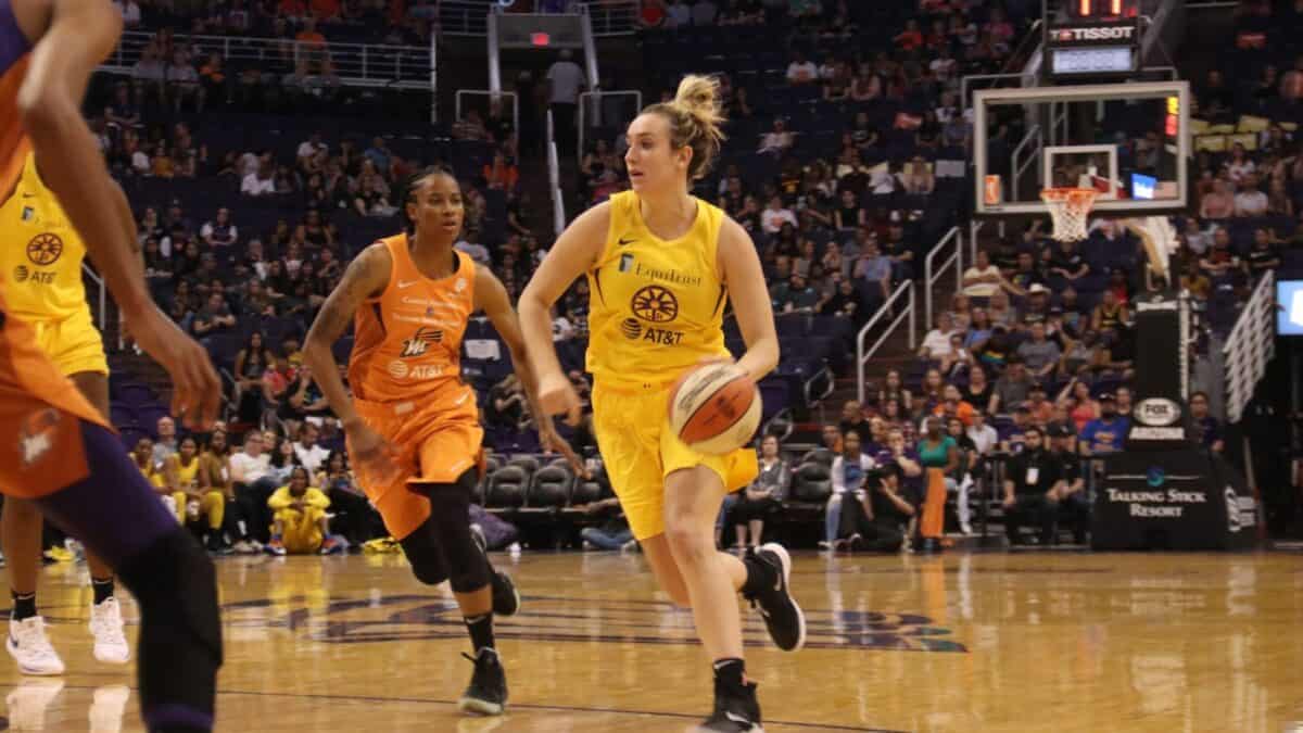 A female basketball player in a yellow uniform is dribbling the ball down the court during a game, with an opponent in an orange uniform close behind. the arena is filled with spectators.