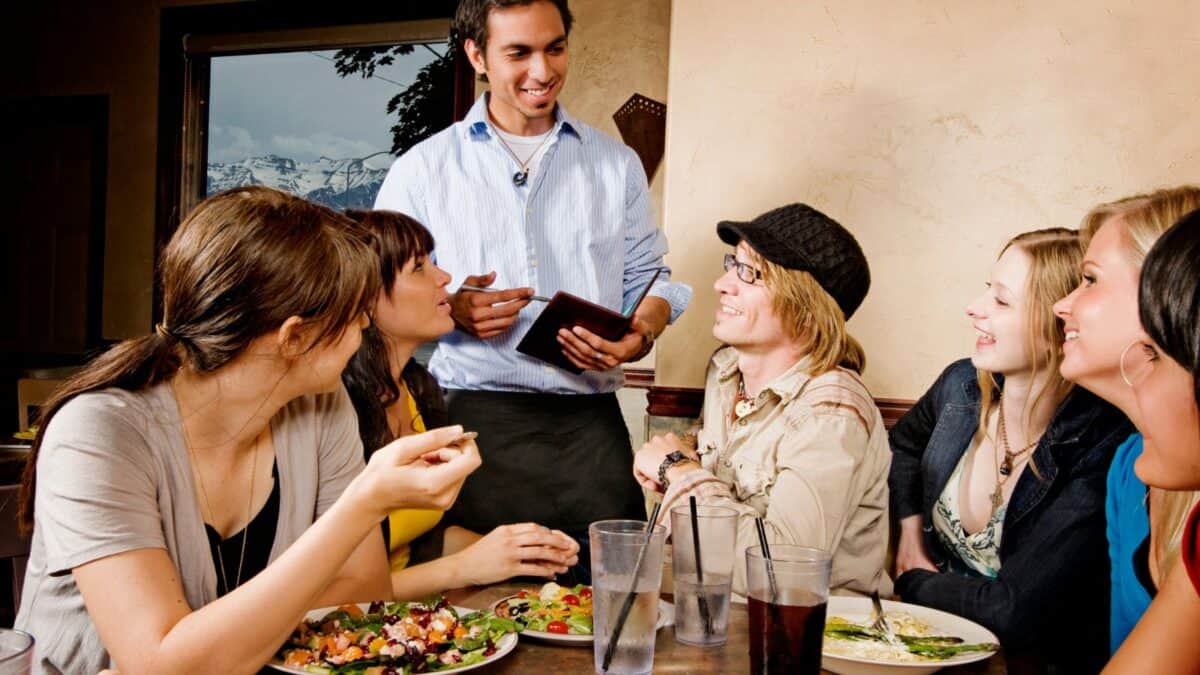 20 Phrases You Should Learn Before You Visit Italy 17 A group of five young adults enjoying a meal at a restaurant, laughing and talking while a smiling waiter takes their order.