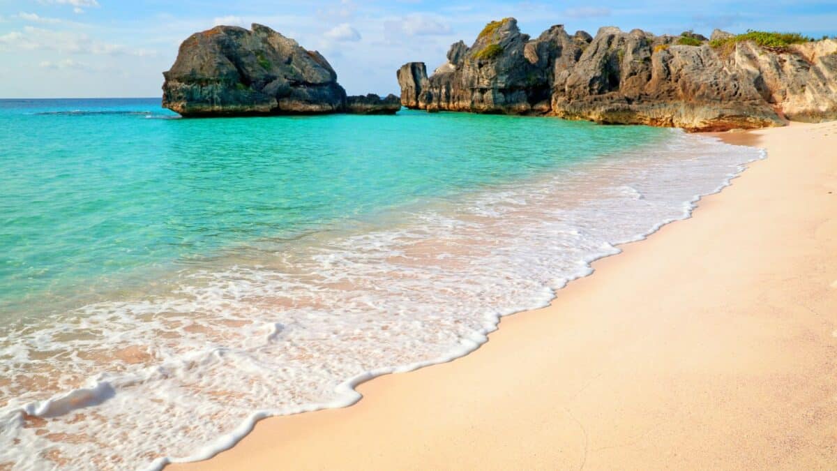Tropical beach with clear turquoise waters gently washing up on a sandy shore, flanked by rugged large rocks under a bright blue sky.