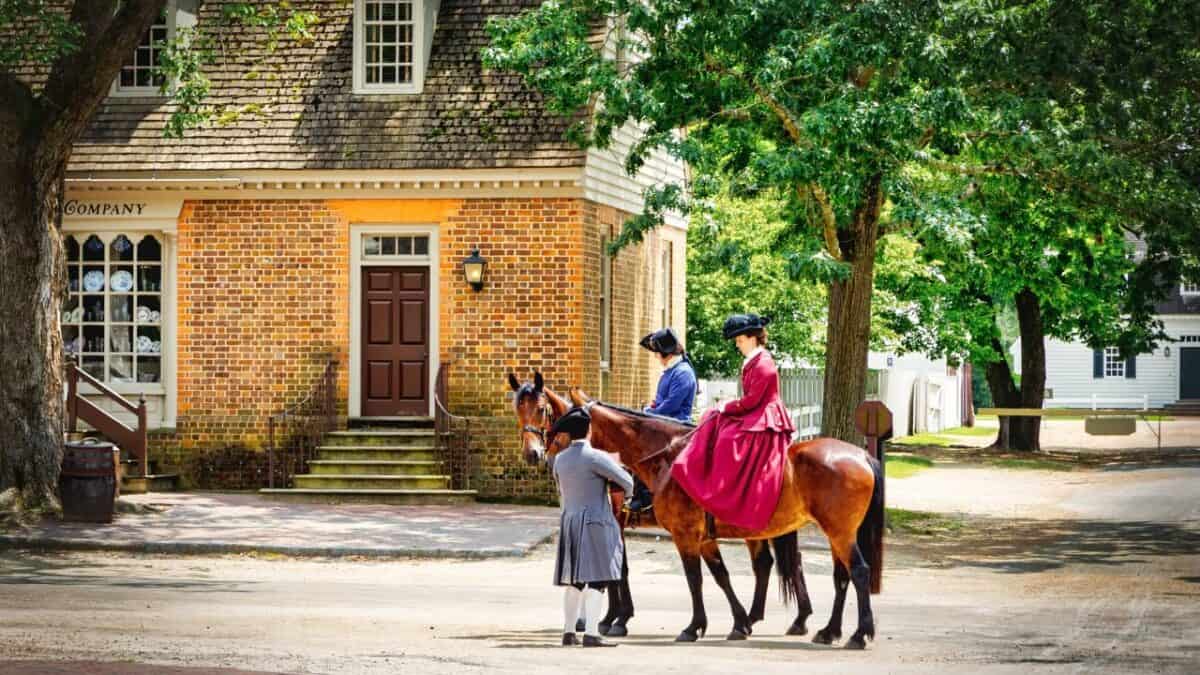 Two people in colonial attire riding horses on a dirt road in front of a historic brick building, with another person in period clothing standing beside them.