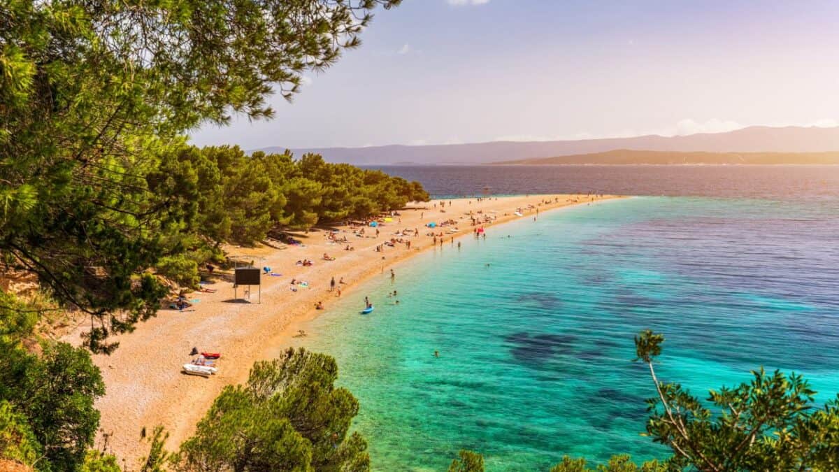 23 Amazing Croatia Landmarks Worth Booking A Trip To See 5 A scenic view of a sunlit beach with clear turquoise waters, surrounded by pine trees, with people enjoying a sunny day.