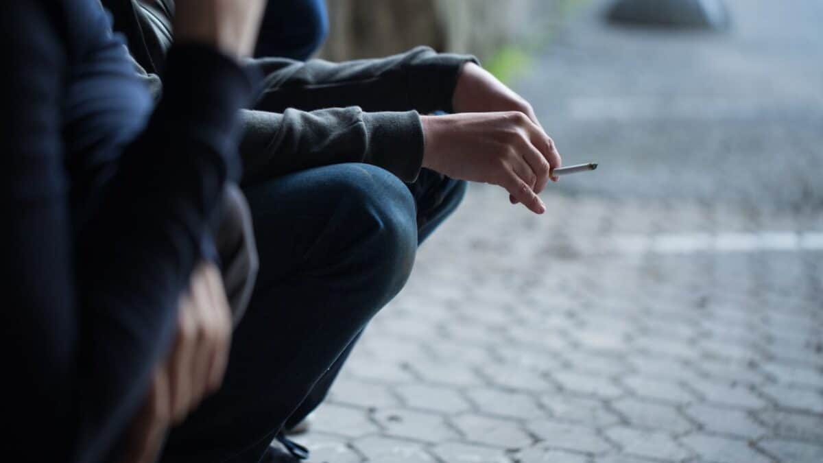 "Close-up of a person seated on a curb, holding a cigarette, with a focus on their hands and the cigarette.