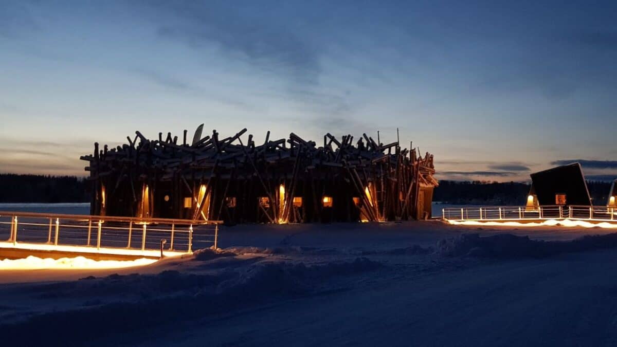 An illuminated, unique wooden structure on a snowy landscape during twilight with a clear sky in the background.