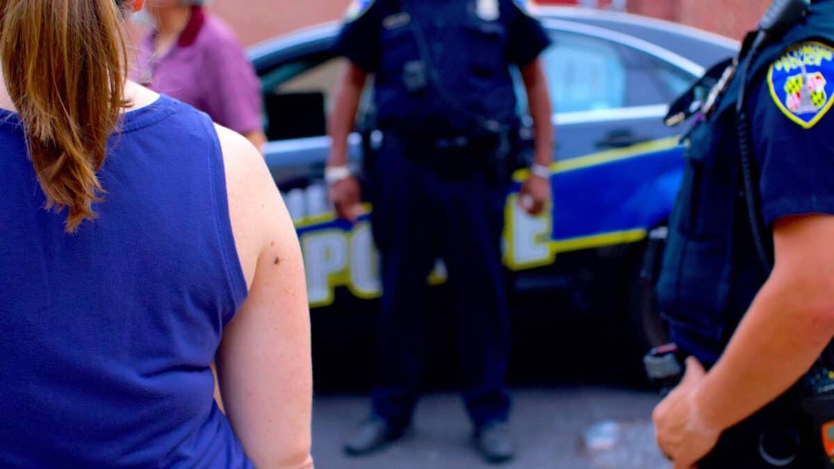 Police officers in uniform standing by a patrol car, engaging with a civilian on a city street, focus on police presence and public interaction.