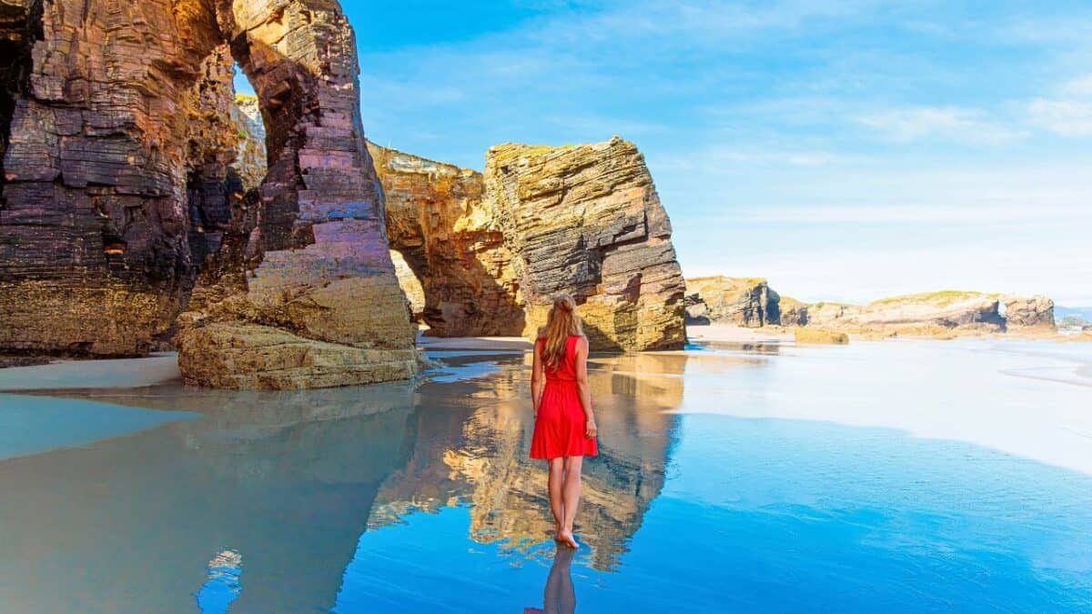 Woman in a red dress standing on a beach, facing large rock formations with arches reflected in a shallow water pool.