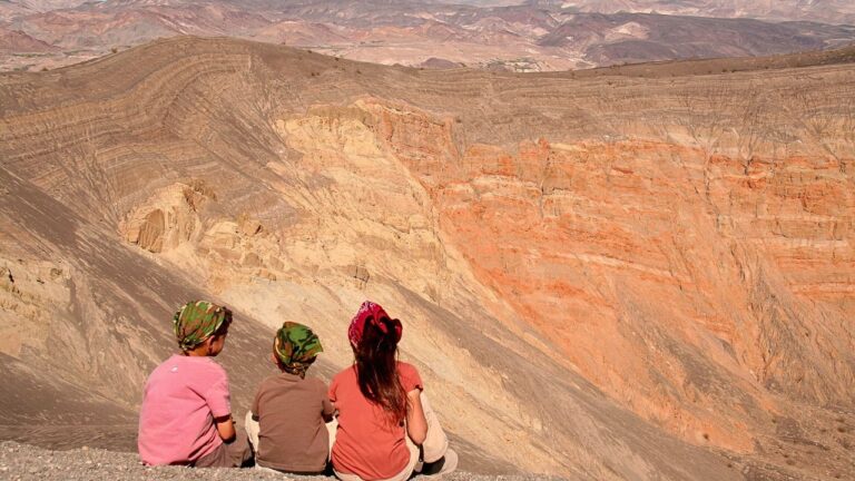Three people, two children and an adult, sit at the edge of a cliff overlooking the colorful, stratified rock formations of Death Valley National Park.