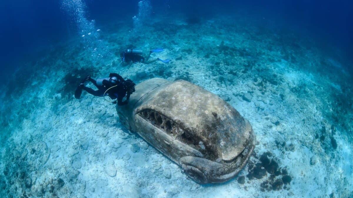 A scuba diver explores near an old, encrusted car submerged on the ocean floor, surrounded by clear blue water and marine life.