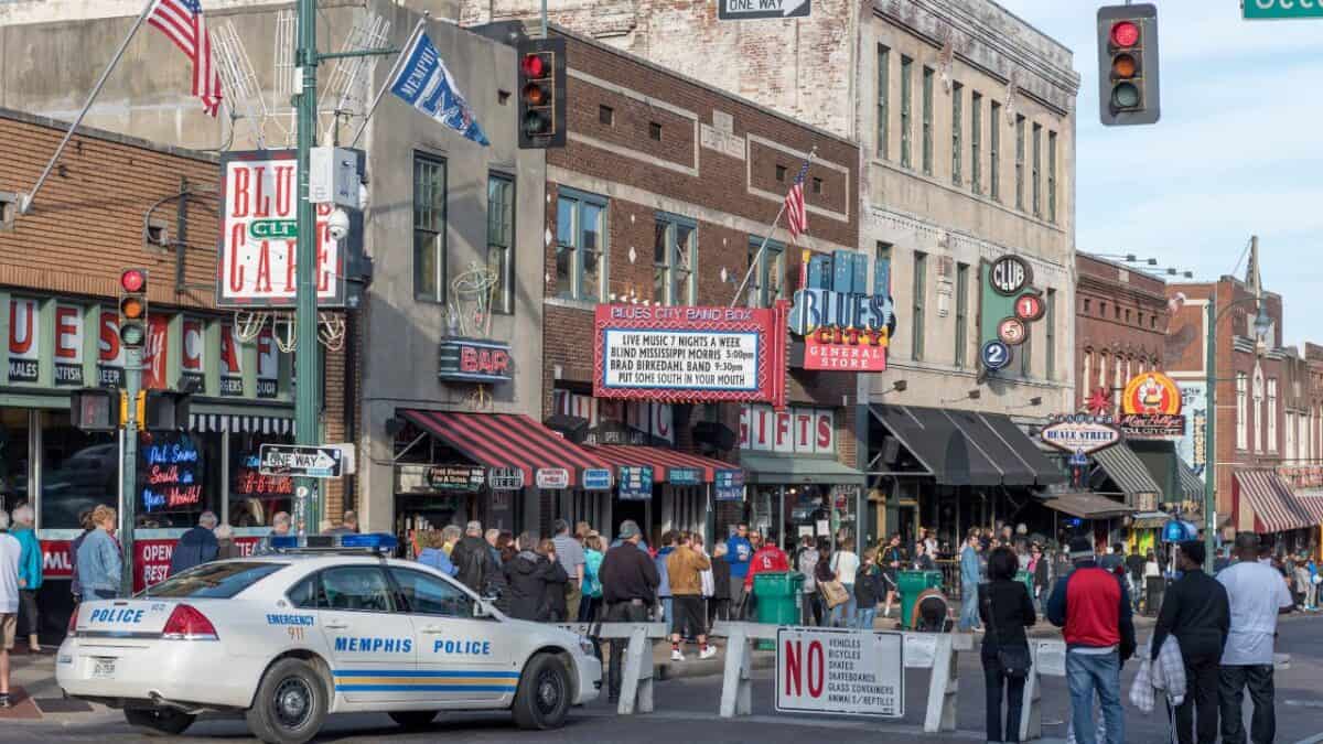 A Memphis Police car in front of a busy street scene with pedestrians and vibrant signs for blues clubs and cafes on Beale Street.