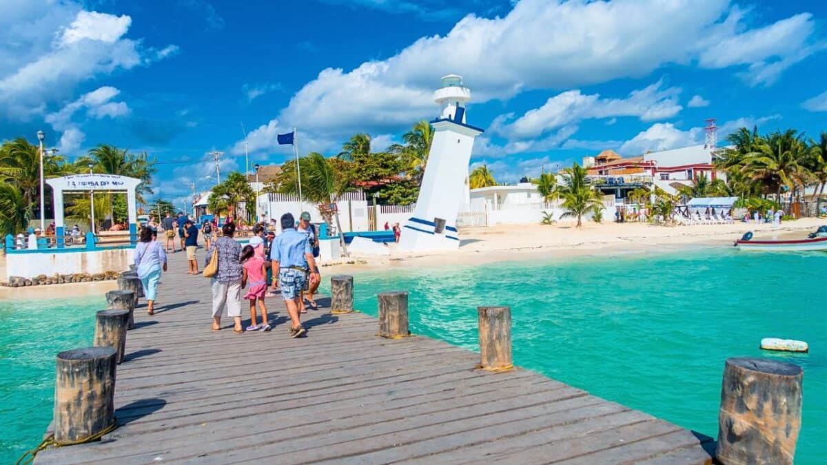 Visitors walking along a wooden pier toward a white lighthouse, with the turquoise sea on one side and a sandy beach with palm trees on the other, under a partly cloudy sky.