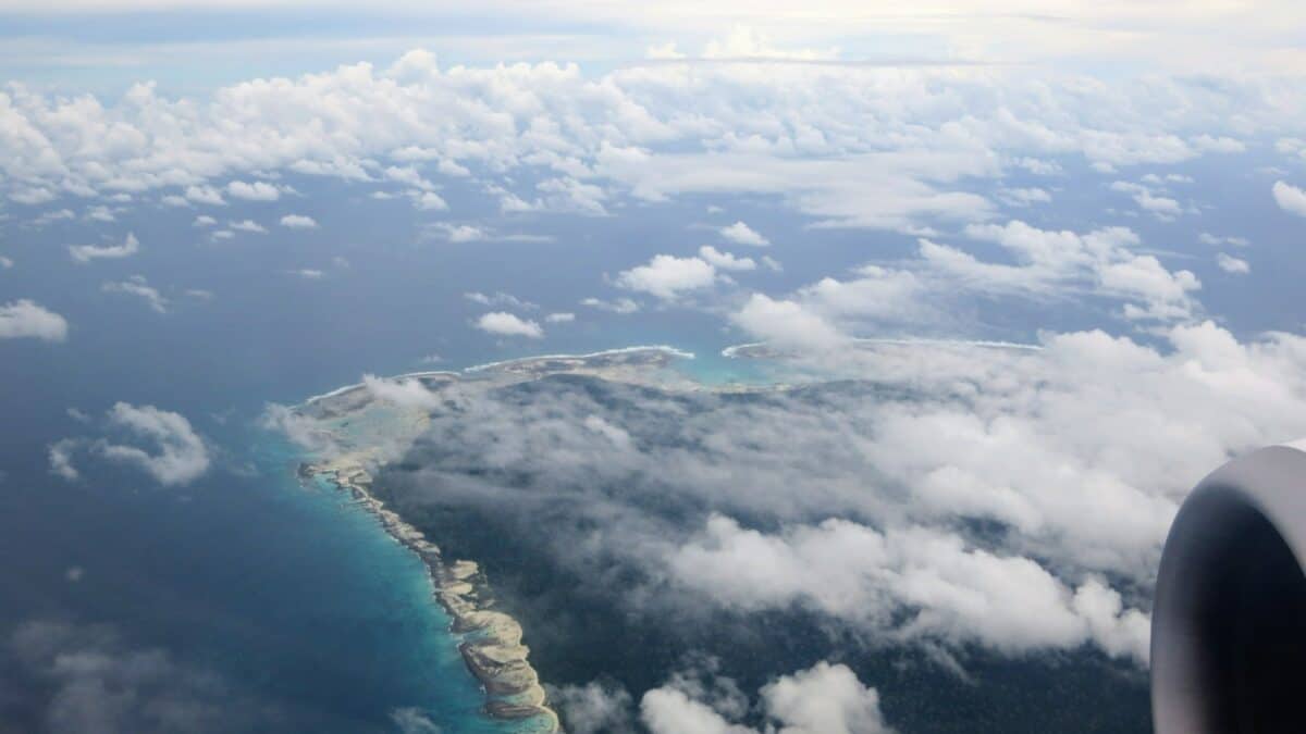 Aerial view from an airplane window showing a stretch of tropical islands surrounded by light blue waters, partially obscured by white, fluffy clouds. Part of the airplane engine is visible on the right.