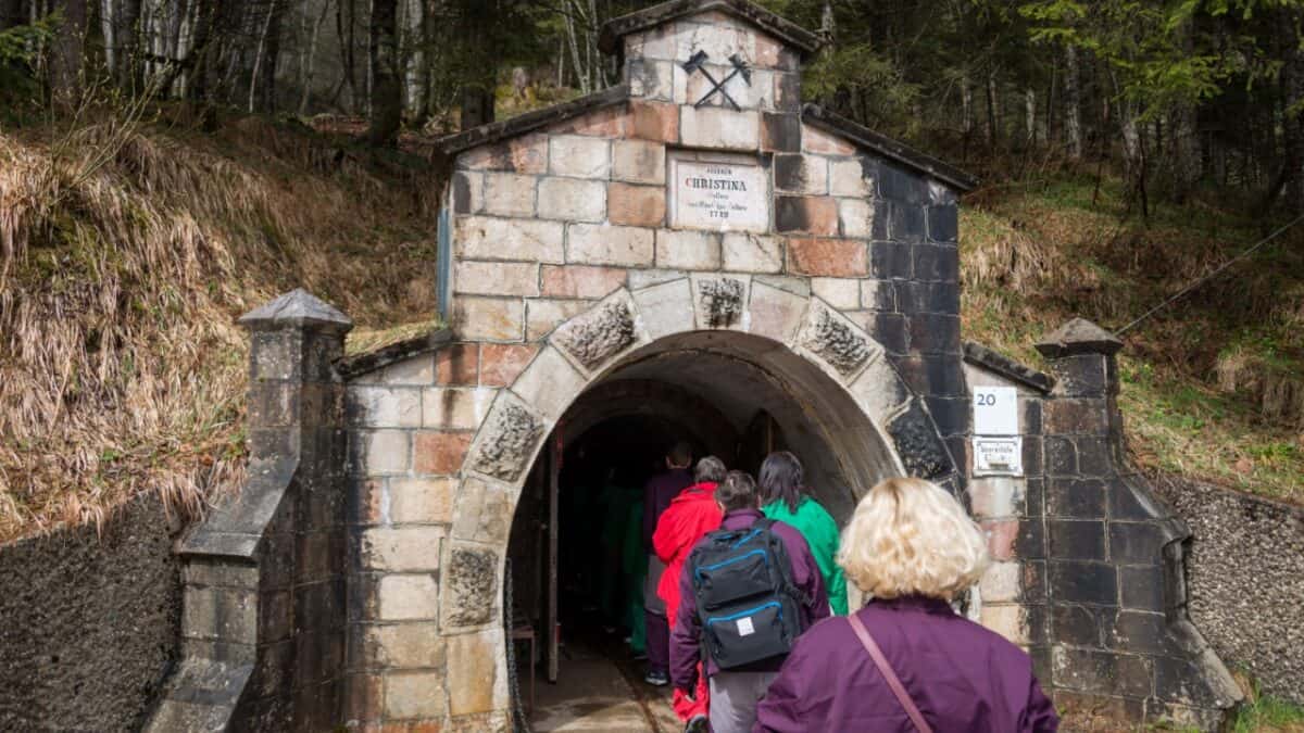 A group of people, some wearing backpacks, are entering a historic stone structure with an arched doorway. The small building is set against a forested hillside. A plaque and mining symbols are visible above the entrance, marking the start of an underground tour in one of the best cities for travel enthusiasts.