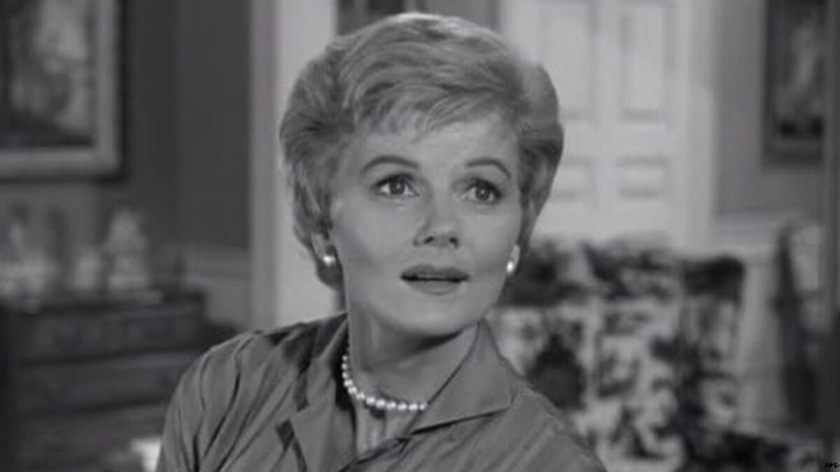 Black and white image of a woman with short blonde hair and pearl earrings, wearing a collared dress and a pearl necklace, looking to her left with a slightly surprised expression.