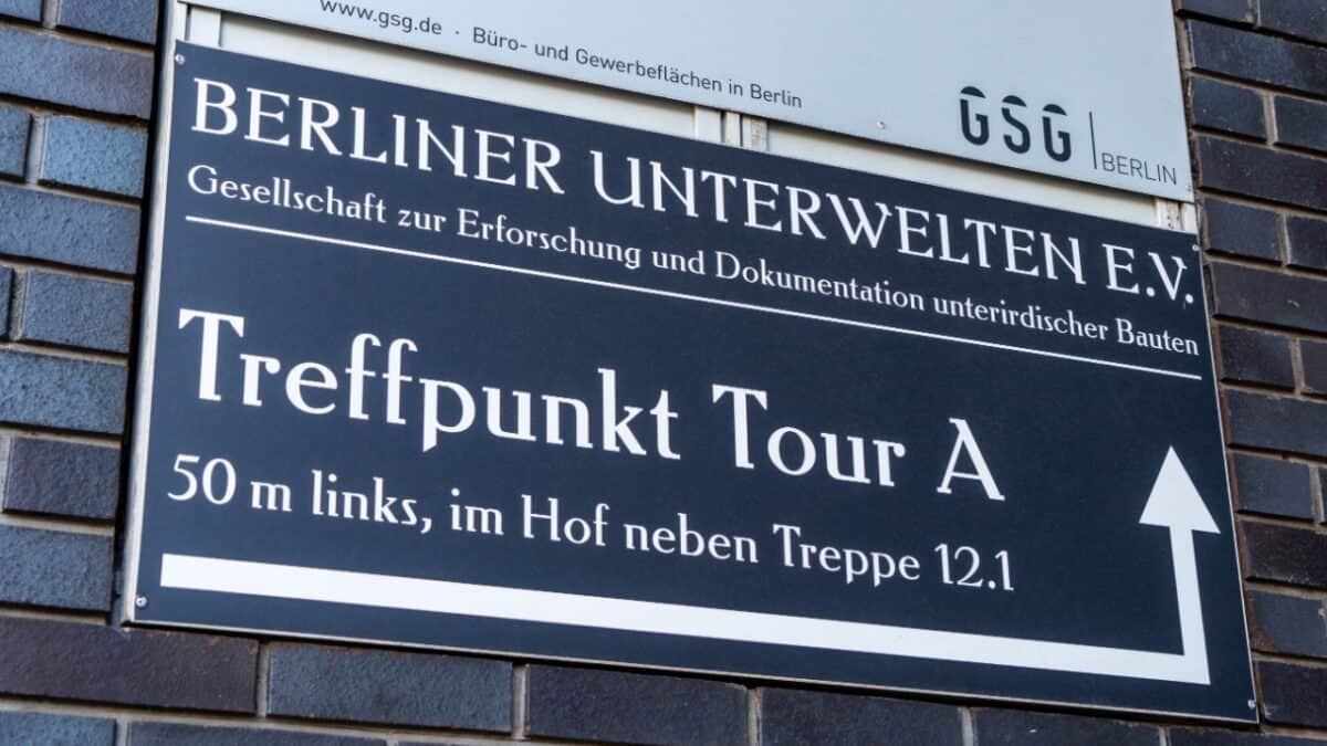 A black and white sign on a brick wall reads, "Berliner Unterwelten E.V, Gesellschaft zur Erforschung und Dokumentation unterirdischer Bauten." It provides directions for "Treffpunkt Tour A," indicating the underground tour is 50 meters to the left, in the courtyard next to stairs 12.1.