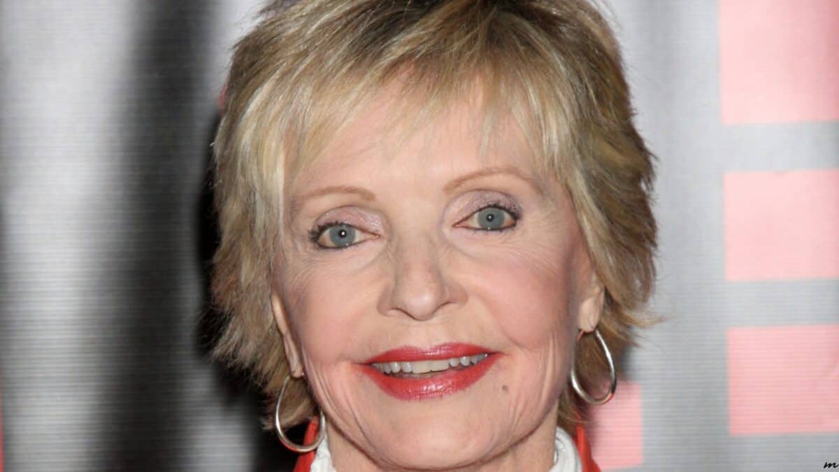 Close-up of a smiling older woman with short blonde hair, wearing red earrings, a white blouse, and a coral cardigan. she's at a red-carpet event with a blurred pink and white backdrop.