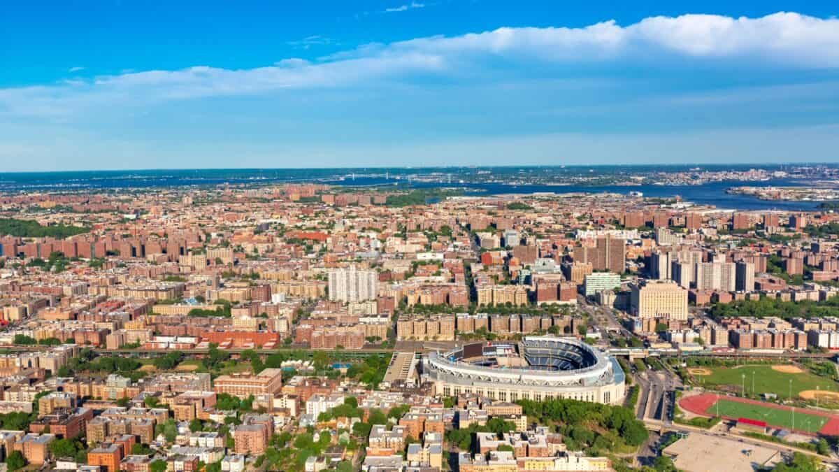 Aerial view of a densely populated urban area with a prominent stadium near the center, surrounded by residential buildings and green spaces, with a river visible in the distance.