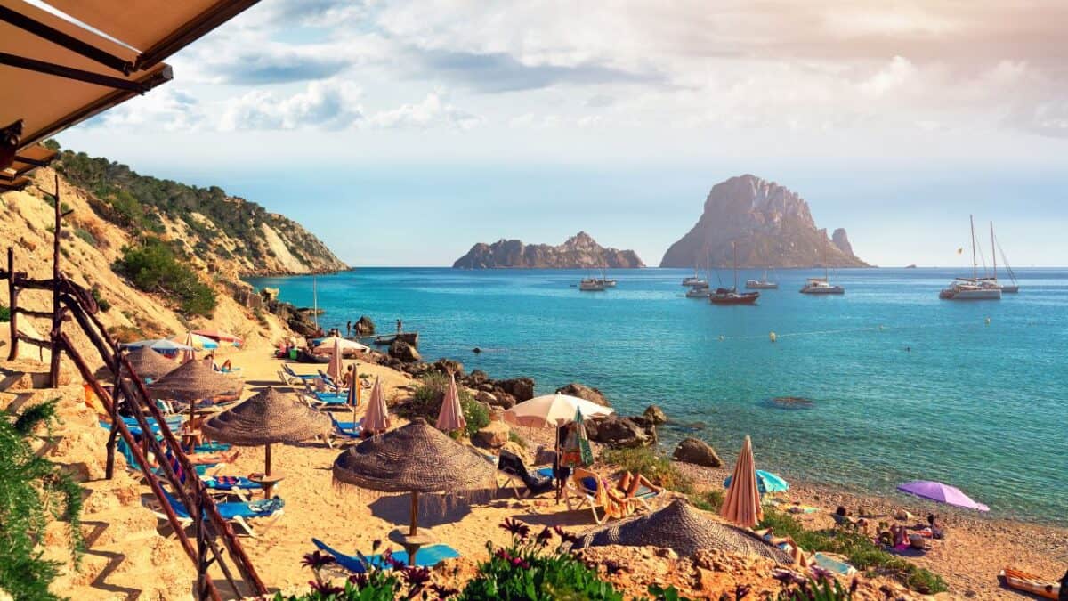 A picturesque beach scene with umbrellas and sun loungers, overlooking clear blue waters and a striking rocky island in the distance. Boats are floating near the shore under a sunny sky.
