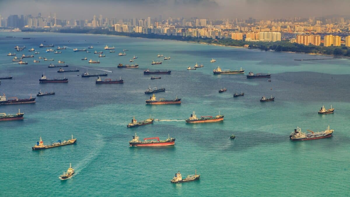 Aerial view of numerous cargo ships and tankers anchored on teal waters near a dense coastal city with high-rise buildings. The skyline appears slightly hazy, and the overall atmosphere suggests busy maritime activity.