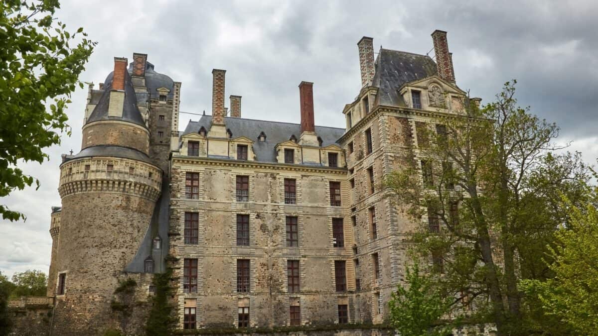 An imposing stone castle with cylindrical towers and multiple chimneys set against a cloudy sky, surrounded by lush greenery.