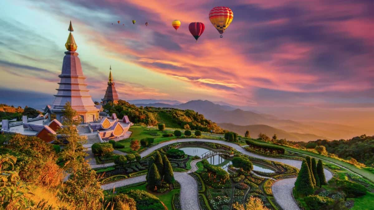 A picturesque landscape featuring two ornate pagodas surrounded by meticulously landscaped gardens. Hot air balloons float in the sky during a colorful sunrise or sunset, with mountains visible in the background—truly one of the best destinations for emotional recovery.