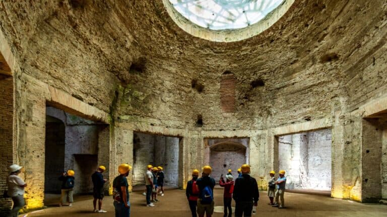 A group of people wearing yellow hard hats stands inside a large, ancient circular structure with high, weathered stone walls. As part of an underground tour in one of the best cities, a circular hole at the top allows light to enter, illuminating the interior. The walls show signs of age and decay.