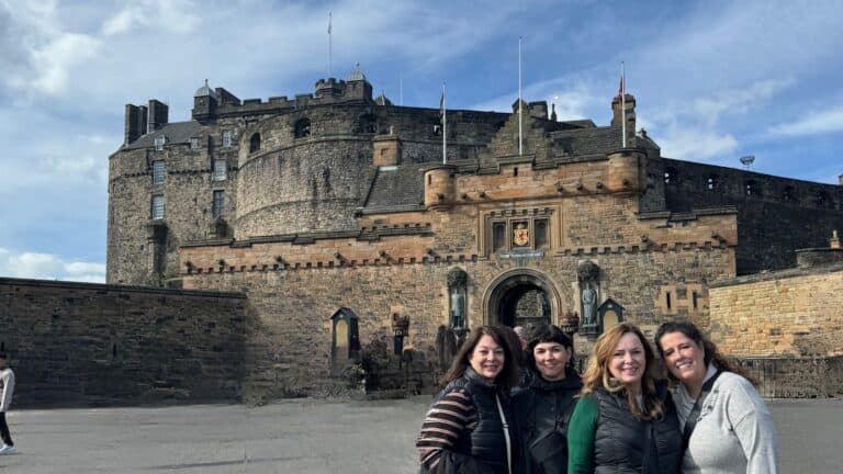 Four women smiling in front of an old castle with a stone facade and a blue sky in the background.