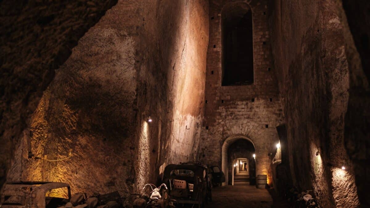 A dimly lit underground passage with tall, rough stone walls. Old machinery and debris are scattered along the floor, leading to an arched stone doorway at the end. Small lights affixed sporadically to the walls cast shadows on uneven surfaces—an eerie backdrop for an underground tour in one of the best cities for travel enthusiasts.