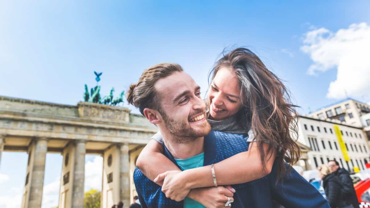 Couple smiling and embracing in front of the Brandenburg Gate on a sunny day.