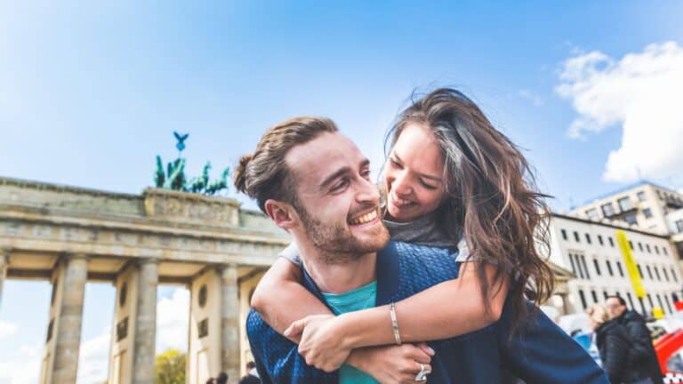 Couple smiling and embracing in front of the Brandenburg Gate on a sunny day.
