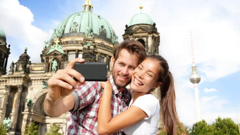A couple takes a selfie in front of a historic building with green domes and a tall tower in the background.