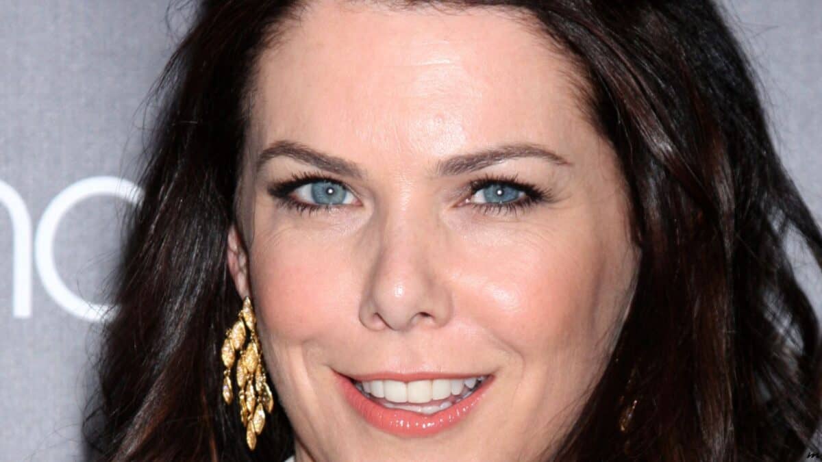 Close-up of a smiling woman with long dark hair at a formal event, featuring striking blue eyes and wearing gold earrings.