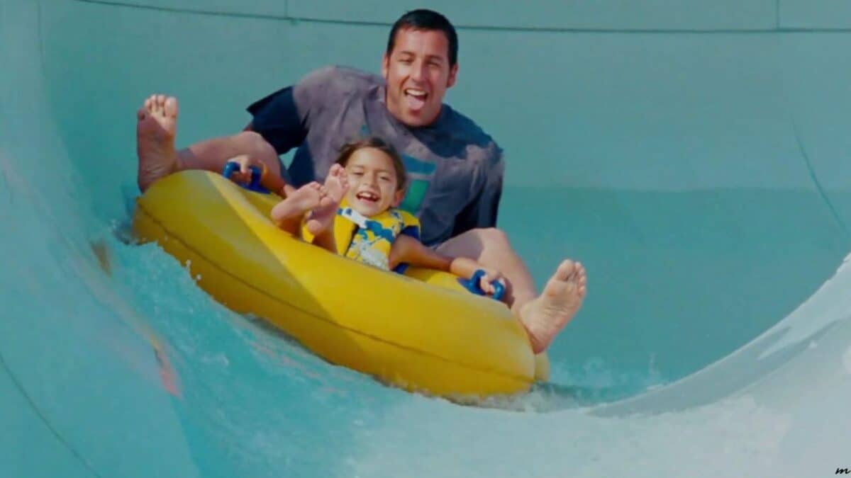 A joyful man and child slide down a water slide in a yellow raft, laughing and with their hands up, in an aqua-colored water park slide.