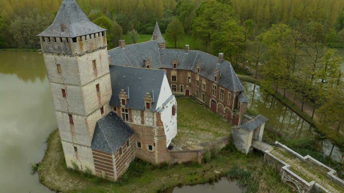 Aerial view of an old european castle surrounded by greenery and a moat, featuring a prominent stone tower and a courtyard.