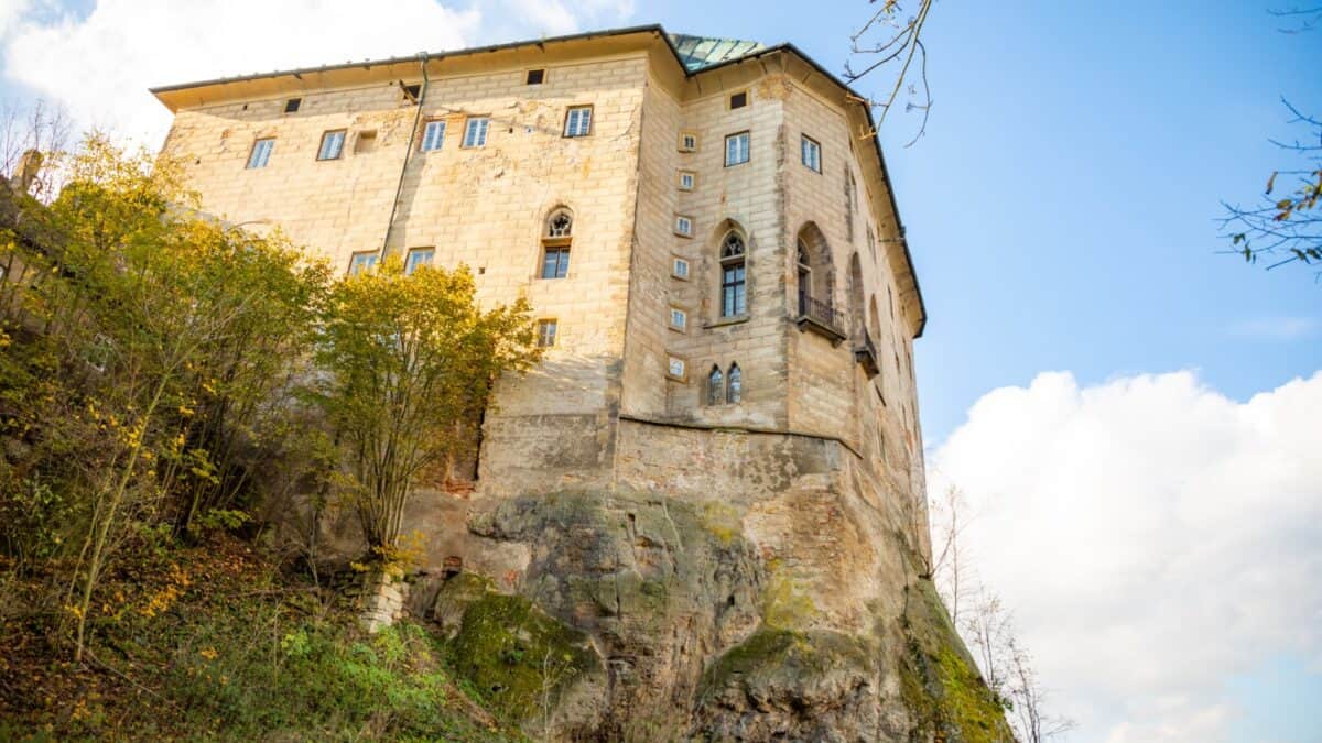 A large, historic stone building perched atop a rugged cliff, surrounded by autumn foliage under a clear blue sky.